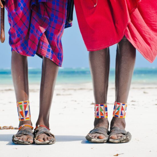 Deux personnes portant des tissus traditionnels rouges et violets, parées de bijoux perlés aux chevilles, debout sur une plage de sable blanc avec la mer turquoise à l’horizon.