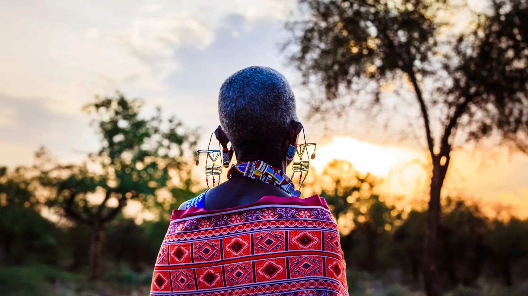 Femme masaï vue de dos, vêtue d’un tissu rouge à motifs et portant des boucles d’oreilles traditionnelles, observant le coucher de soleil en Tanzanie.