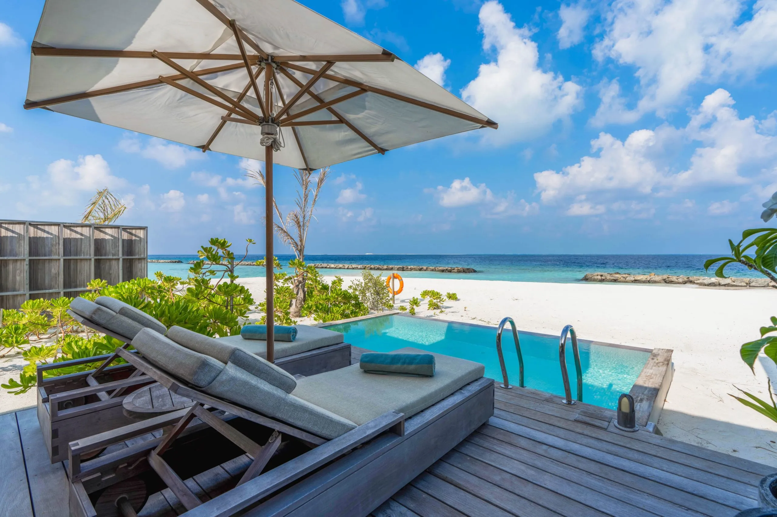 Plage paradisiaque avec transats et piscine, sous un ciel lumineux et des eaux turquoise.