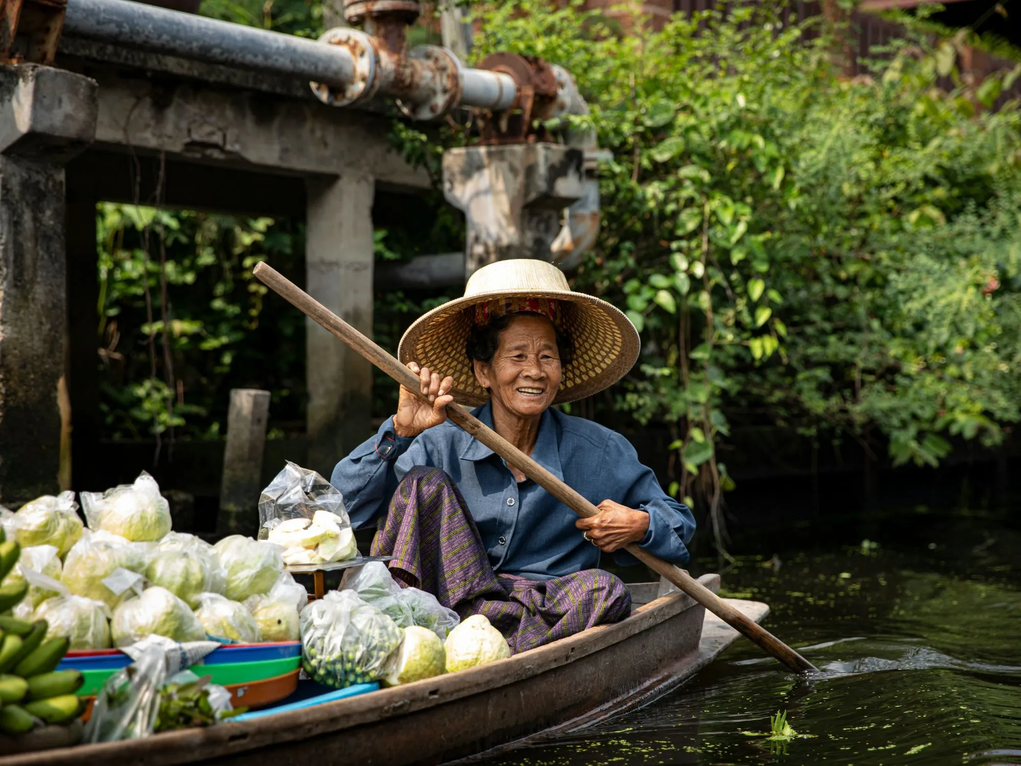 Femme souriante sur une barque, entourée de légumes frais dans un cadre verdoyant et paisible.