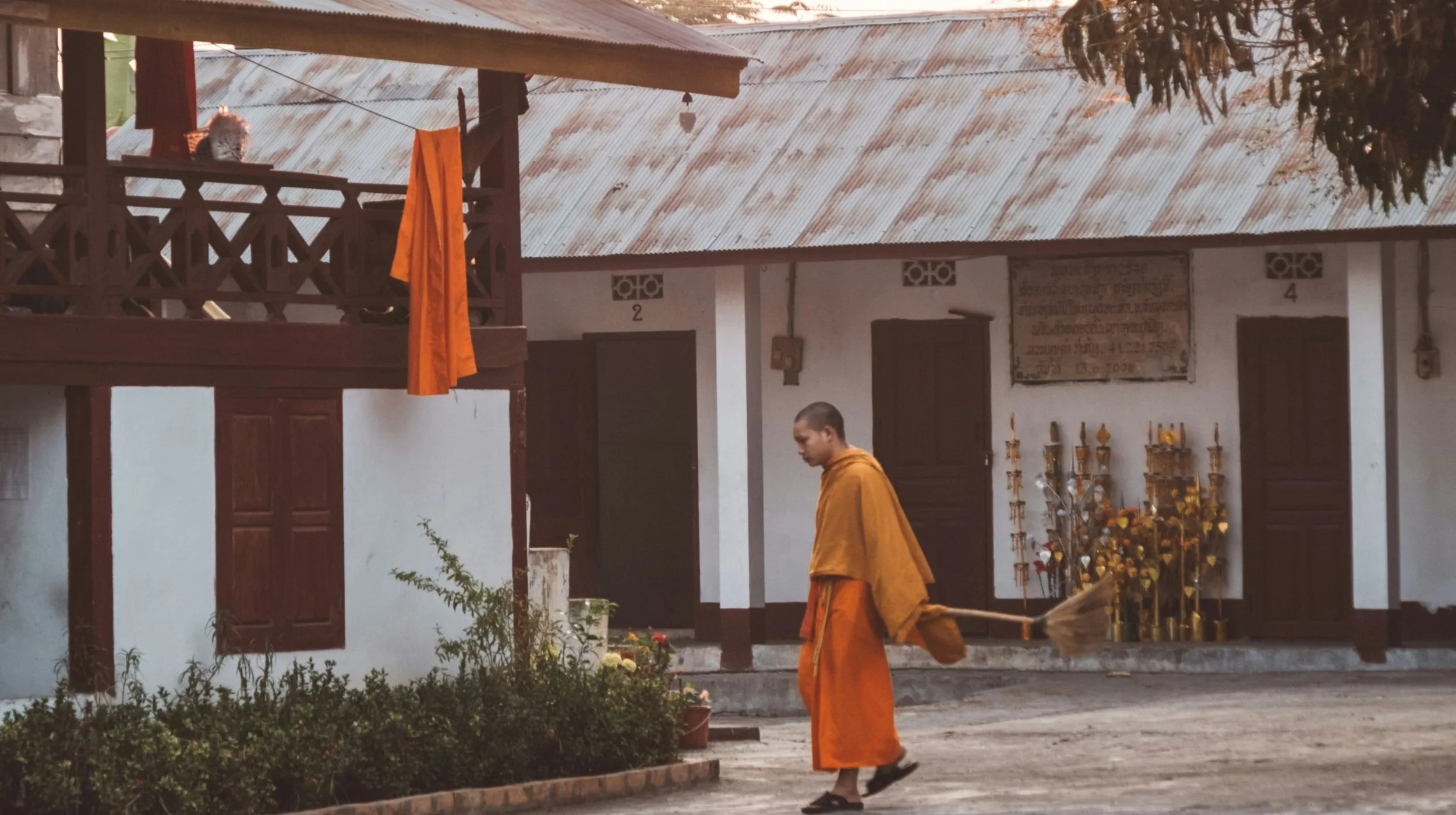 Moine en robe orange dans une cour paisible, entouré de bâtiments traditionnels et de végétation luxuriante.