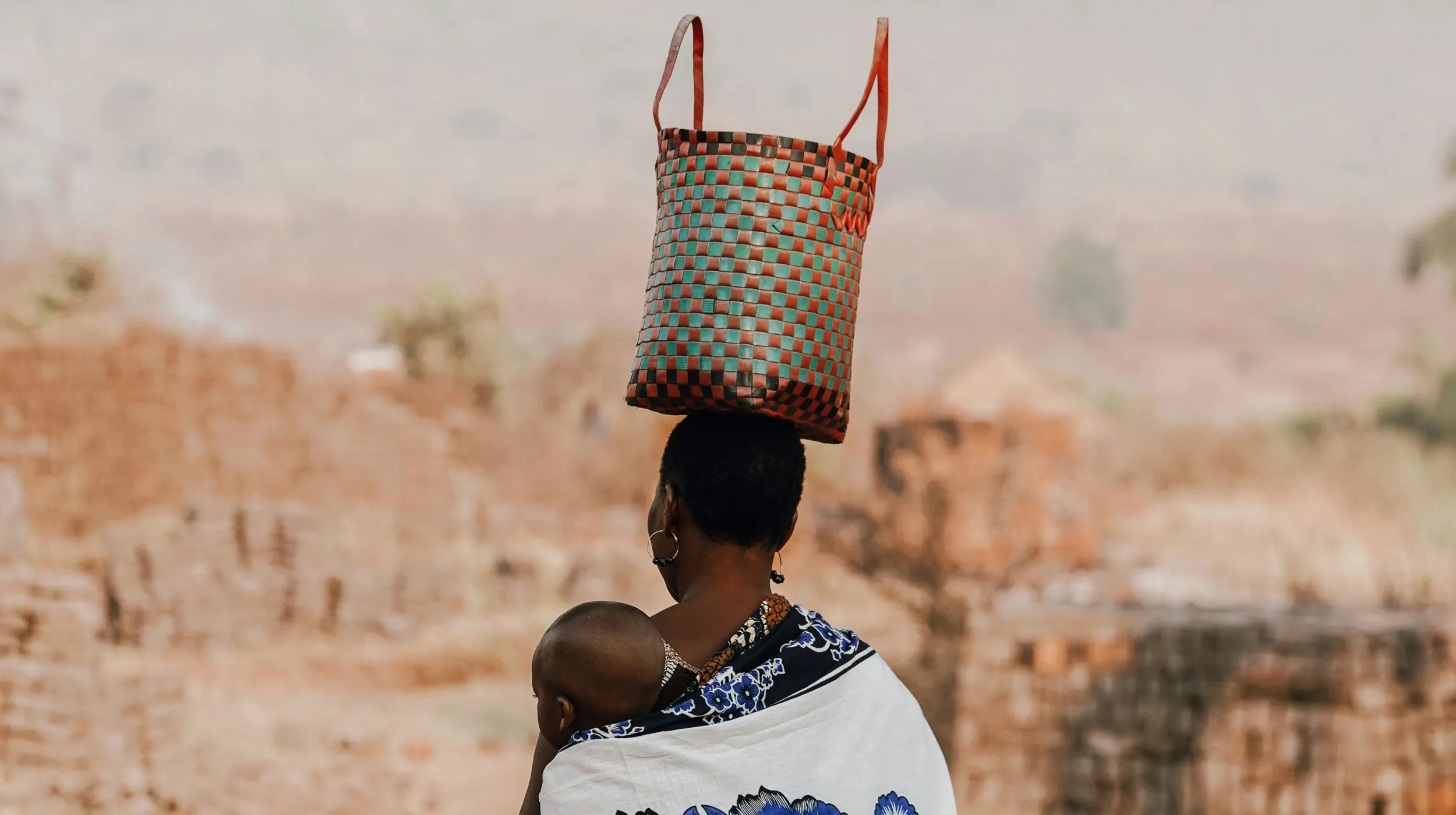 Femme portant un enfant et un panier coloré sur la tête, dans un paysage rural apaisant.
