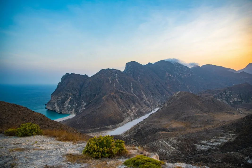 Paysage côtier d’Oman au coucher du soleil, avec des montagnes arides plongeant dans la mer turquoise et une vallée rocheuse serpentine au premier plan.